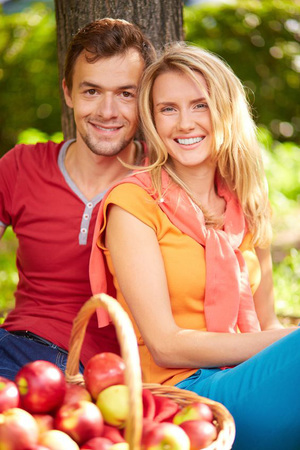 A man and a woman lean against a tree with a basket of apples